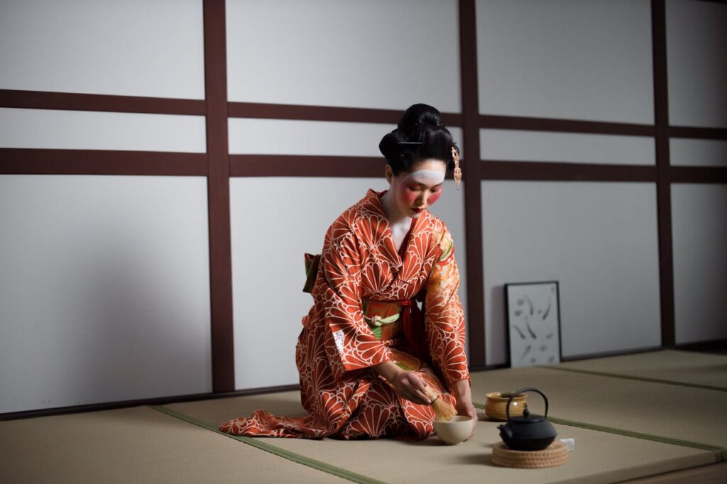 Is Matcha the New Perfume Trend? 19 A woman in a bright orange kimono making matcha tea with a bamboo whisk during a traditional Japanese tea ceremony in a simple tatami room.