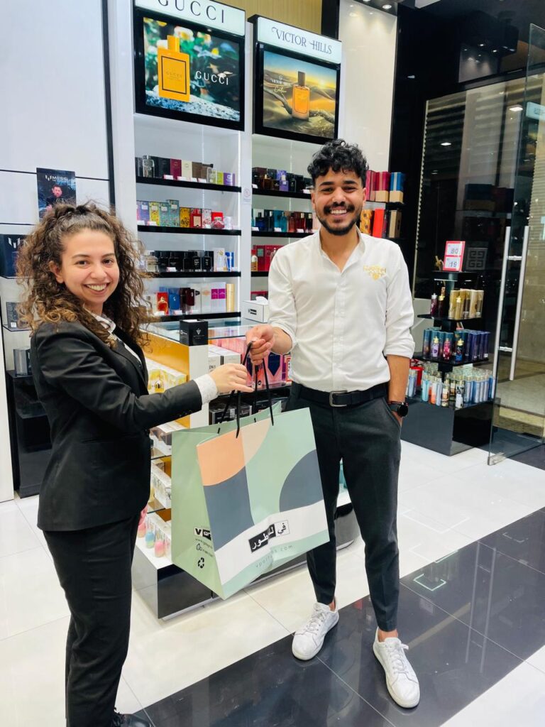 In this image a smiling man and woman stand inside a perfume store, exchanging a shopping bag. The woman, dressed in a black blazer and white shirt, extends the bag toward the man, who is wearing a white shirt with a golden logo, black trousers, and white sneakers. The store shelves behind them are filled with luxury perfume brands like Gucci and Victor Hills, with a sleek, modern design featuring white and black glossy tiles.