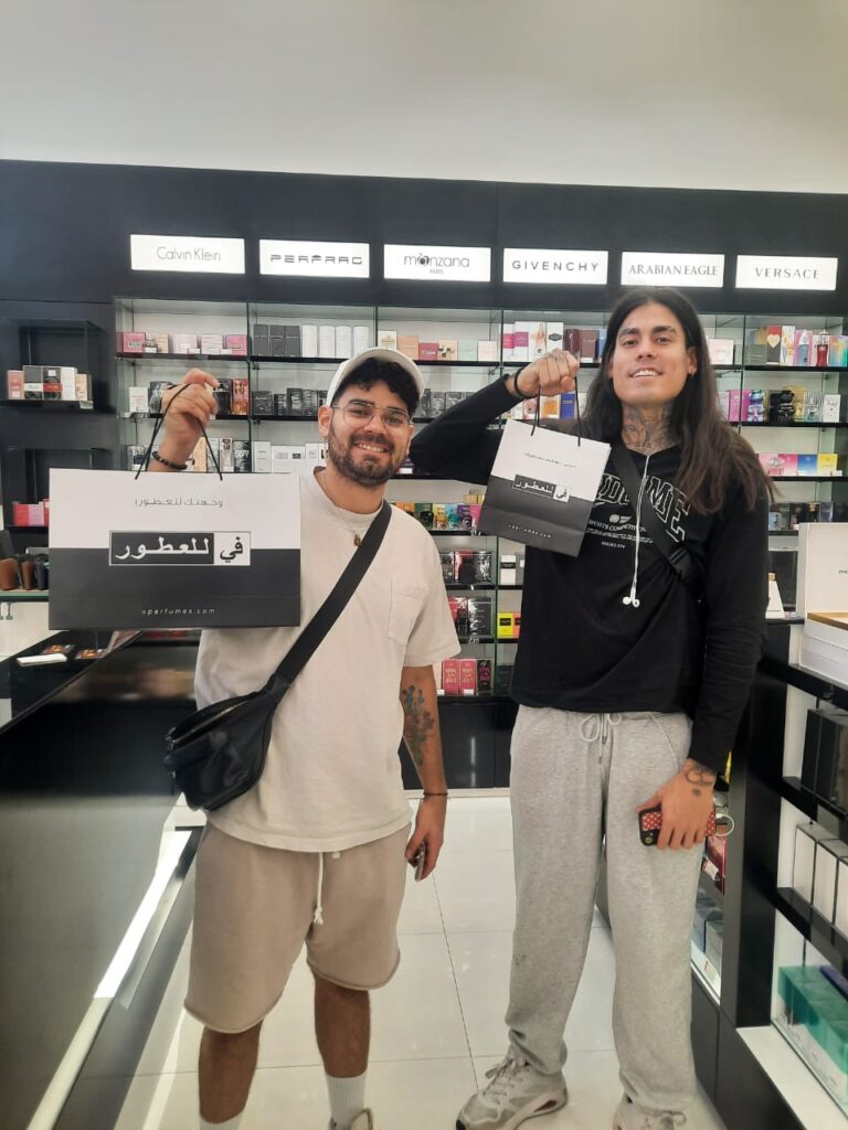 In this image two young men with light to medium skin tones are standing inside a V Perfumes store, smiling and holding up branded shopping bags containing their winnings from the Youm Al-Ottoor event. The store shelves behind them display various perfume brands, including Calvin Klein, Givenchy, and Versace. The man on the left wears a white t-shirt, beige shorts, a white cap, glasses, and a black crossbody bag. The man on the right wears a black long-sleeve shirt, gray sweatpants, white sneakers, and has long dark hair. He also has tattoos on his arms and is holding a phone. The shopping bags they hold feature Arabic text and the website "vperfumes.com". The store has a sleek black and white interior with a glossy floor reflecting the bright lighting.